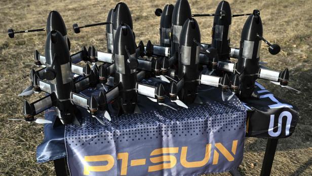 Mehrere schwarze Drohnen mit Propellern stehen auf einem Tisch im Freien auf einer blauen Decke mit gelber Aufschrift.