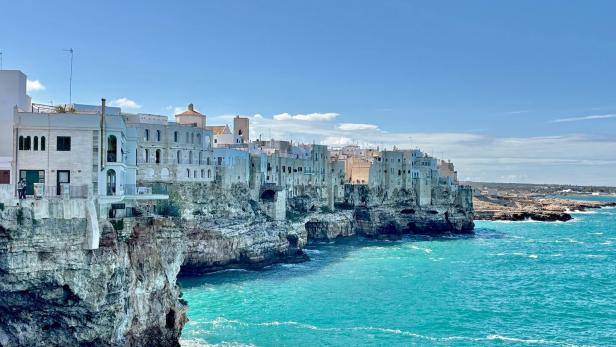Weiße Häuser thronen auf Klippen über dem türkisfarbenen Meer unter blauem Himmel.