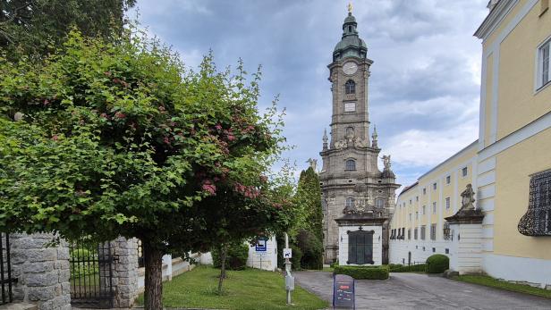 Ein Kirchturm mit Uhr steht neben einem gelben Gebäude und einem Baum mit rosa Blüten.