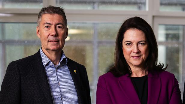 Ein Mann im Sakko und eine Frau im lila Blazer stehen nebeneinander vor einem Fenster.