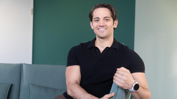 Ein Mann mit dunklem Haar und schwarzem Polohemd sitzt lächelnd auf einem Sofa vor einer grünen Wand.