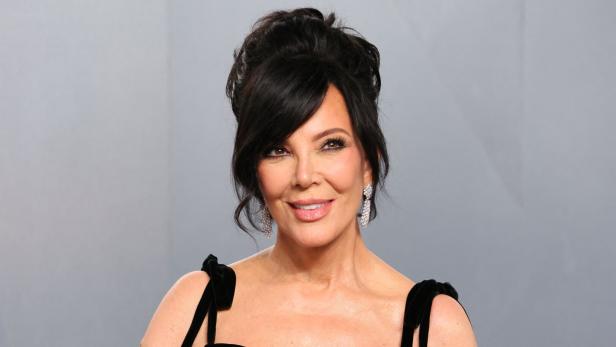 Kris Jenner trägt ein schwarzes Kleid und auffällige Ohrringe vor einem grauen Hintergrund.