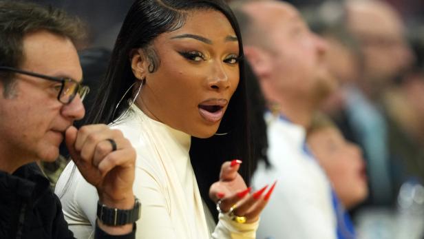 Megan Thee Stallion schaut mit offenem Mund, roten Nägeln und großen Ohrringen überrascht.