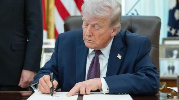 Donald Trump unterzeichnet ein Dokument mit einem Stift vor einer US-Flagge.