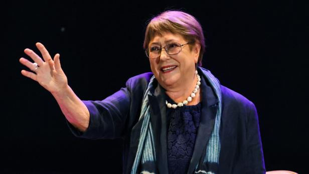Michelle Bachelet lächelt und winkt mit erhobener Hand vor dunklem Hintergrund.
