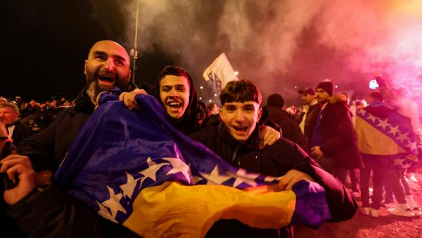 Drei jubelnde Menschen halten eine bosnische Flagge, umgeben von einer feiernden Menschenmenge bei Nacht und Rauch.