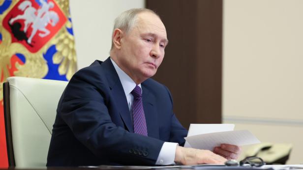 Putin sitzt am Tisch und liest ein Dokument vor einer russischen Flagge.