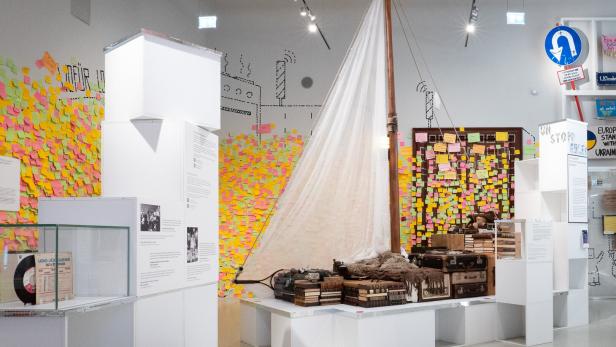 Ein Segelboot mit Büchern, Koffern und Alltagsgegenständen steht in einem Ausstellungsraum, die Wände sind mit bunten Notizzetteln bedeckt.