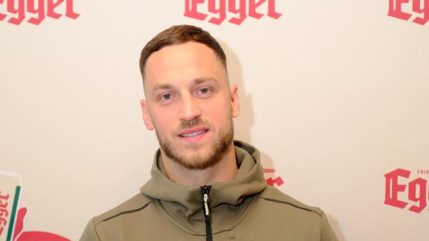 Ein Mann in olivgrüner Jacke hält eine Getränkedose mit rotem Etikett vor einer Wand mit Egger-Logos.