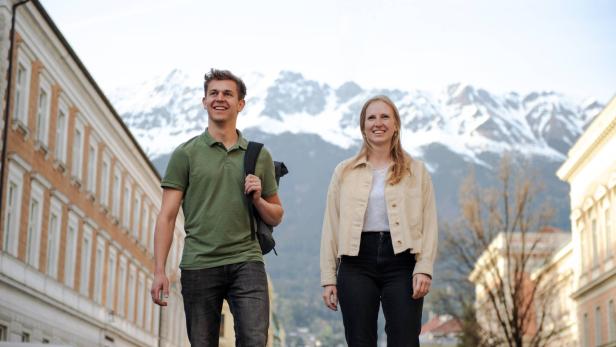 Zwei junge Menschen gehen lächelnd durch eine Stadt mit schneebedeckten Bergen im Hintergrund.