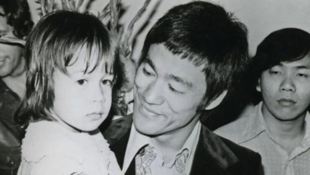 Bruce Lee hält ein kleines Mädchen im Arm, während andere Personen im Hintergrund stehen.