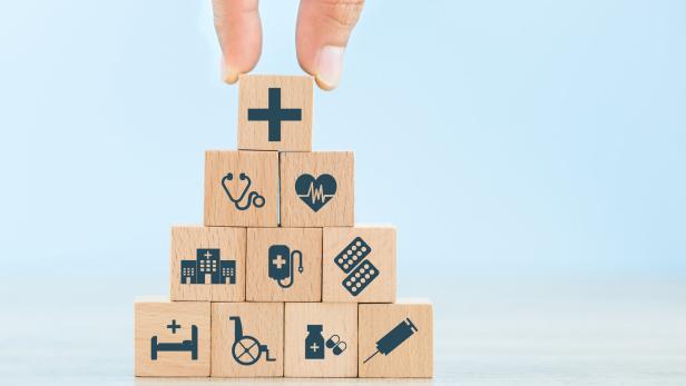 Das Foto zeigt zu einem Turm aufgestapelte Holzwürfel mit kleinen graphischen Zeichen auf der Vorderseite, die symbolhaft für das Gesundheitssystem stehen wie etwa ein Stethoskop, eine EKG-Linie oder eine Spritze.