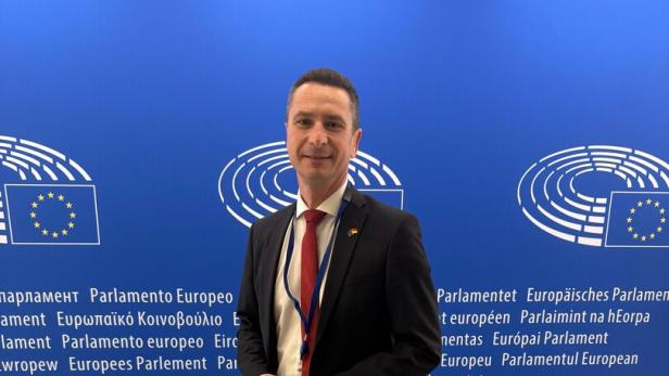 Ein Mann im Anzug steht vor einer blauen Wand mit Logos und Schriftzügen des Europäischen Parlaments.