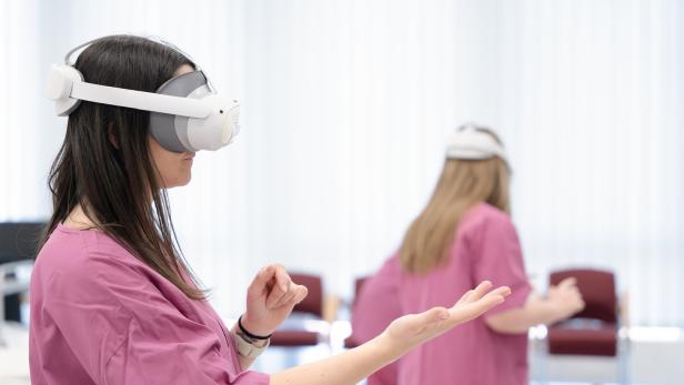 Zwei Personen in rosa Kitteln tragen Virtual-Reality-Headsets und führen gestikulierende Bewegungen in einem hellen Raum aus.