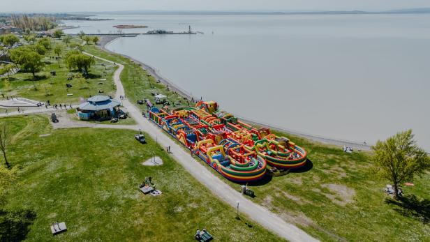 Großer bunter Hüpfburgen-Parcours am Ufer eines Sees, umgeben von Wiese, Wegen und Bäumen mit Menschen.