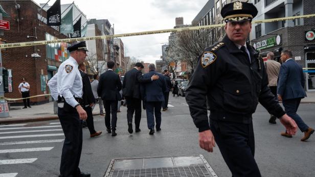 Mehrere Polizisten und Männer in Anzügen stehen und gehen auf einer abgesperrten Straße mit gelbem Absperrband.