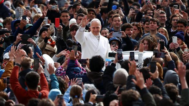 Ein Mann in weißer Kleidung winkt einer großen, jubelnden Menschenmenge, viele halten ihre Handys hoch.