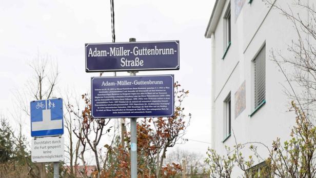 Drei Personen und ein Hund stehen in der Adam-Müller-Guttenbrunn-Straße vor einem Straßenschild mit Informationstafel.