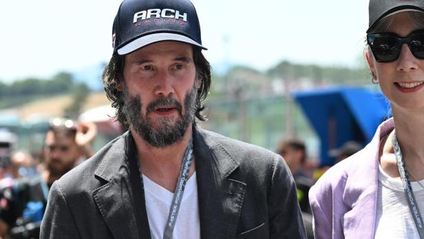 Keanu Reeves mit Bart und Kappe "Arch Racing" trägt ein Jackett.