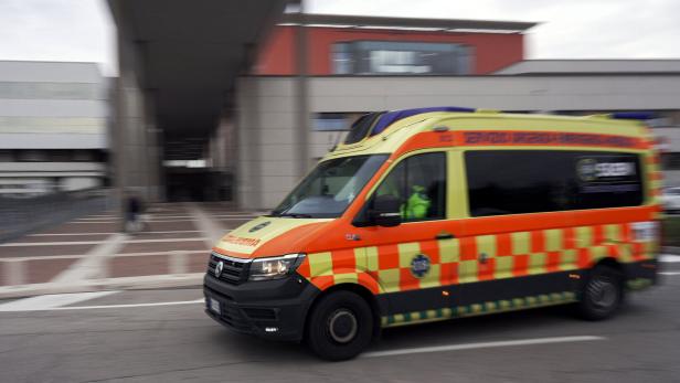Ein gelb-orangefarbener Rettungswagen fährt schnell auf einer Straße vor modernen Gebäuden.