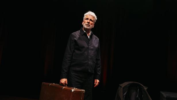 Dirk Stermann hält einen alten Koffer vor schwarzem Hintergrund.