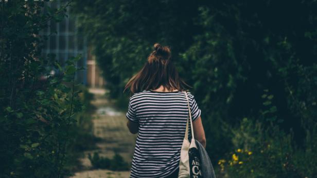 Mädchen mit gestreiftem Shirt und Tasche geht einen von Büschen gesäumten Weg entlang.