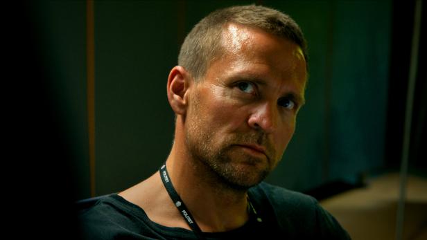 Ein Mann mit kurzem Haar und Bart trägt ein schwarzes T-Shirt und einen Ausweis mit der Aufschrift „Politet“ um den Hals.