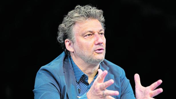 Ein Mann mit lockigen Haaren und Bart gestikuliert mit beiden Händen und trägt ein blaues Sakko über einem gemusterten Hemd.