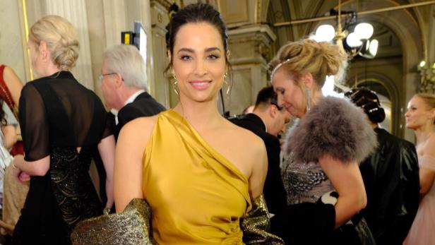 Amira Aly in einem goldenen, schulterfreien Kleid posiert auf einem roten Teppich.