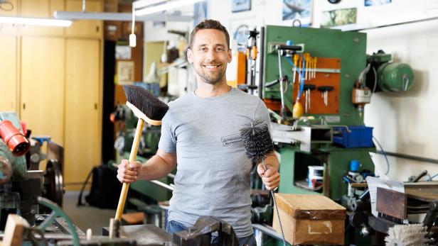 Ein Mann steht in einer Werkstatt und hält einen Besen und eine Bürste in den Händen, umgeben von Maschinen und Werkzeugen.