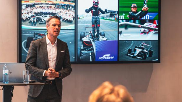 Ein Mann im Anzug hält einen Vortrag vor einer Leinwand mit Motorsport-Bildern und der Überschrift „Erste Punkte und Gesamtführung“.