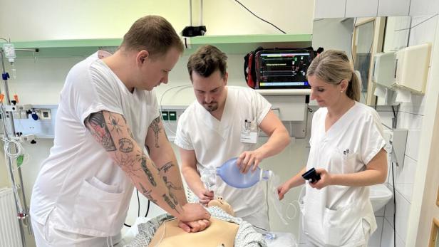 Drei Personen in weißer Kleidung üben an einer Reanimationspuppe Herz-Lungen-Wiederbelebung.