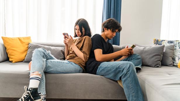 Zwei Jugendliche sitzen Rücken an Rücken auf einem Sofa und schauen konzentriert auf ihre Smartphones.