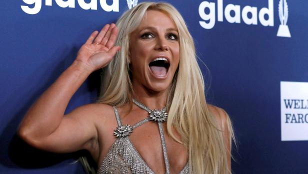 Britney Spears mit erhobener Hand und offenem Mund vor einem GLAAD-Hintergrund.