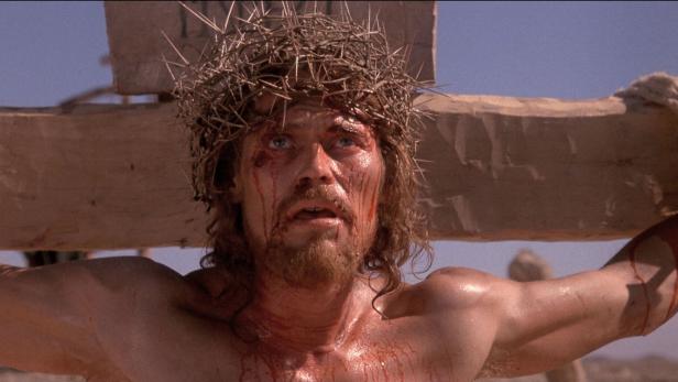 Ein Mann mit Dornenkrone und blutverschmiertem Gesicht hängt mit ausgebreiteten Armen an einem Holzbalken.