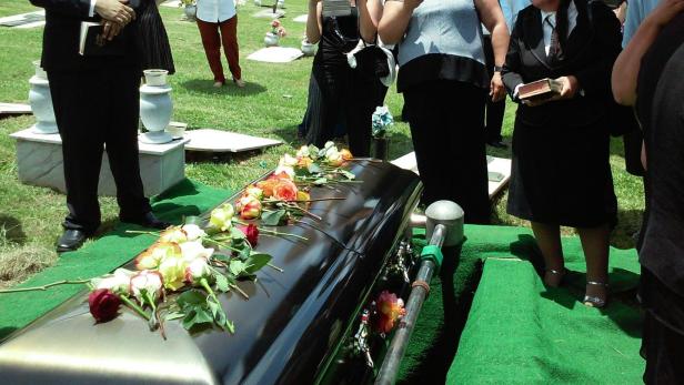 Trauergäste stehen um einen mit Blumen geschmückten Sarg auf einem Friedhof.