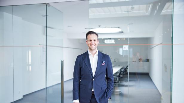 Ein Mann im Anzug steht lächelnd in einem modernen, hellen Büro mit Glastüren und Konferenztisch.
