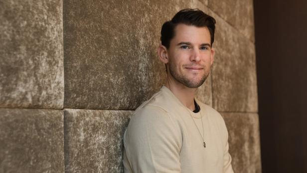 Ein junger Mann mit kurzem dunklem Haar sitzt entspannt vor einer gepolsterten, beigen Wand und lächelt leicht.