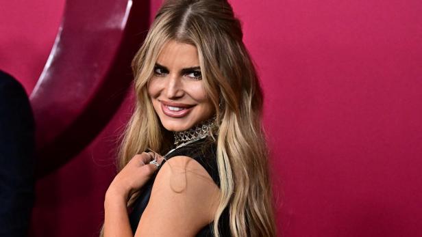 Jessica Simpson mit blonden Haaren und einem schwarzen Kleid lächelt vor einem pinkfarbenen Hintergrund.