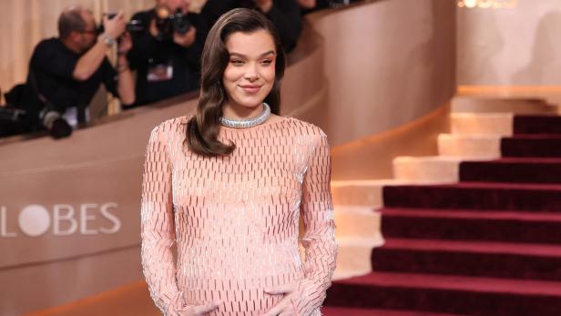 Hailee Steinfeld in einem langen, hellrosa Kleid mit silbernen Akzenten posiert lächelnd vor einer Treppe.
