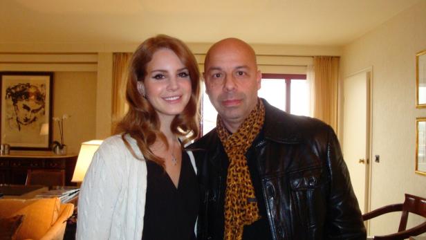 Eine Frau mit langen, braunen Haaren steht neben einem Mann mit Glatze und Lederjacke in einem gemütlich eingerichteten Raum.