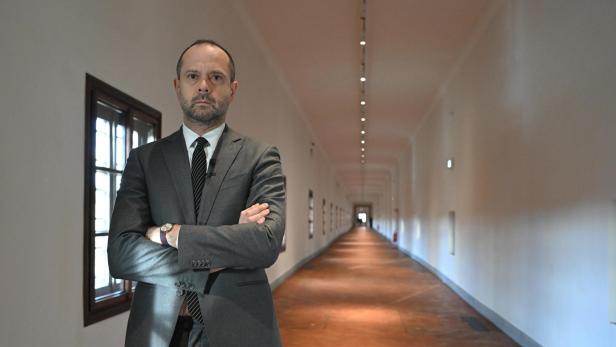 Ein Mann im Anzug steht mit verschränkten Armen in einem langen, hellen Flur mit Fenstern und Bildern an den Wänden.