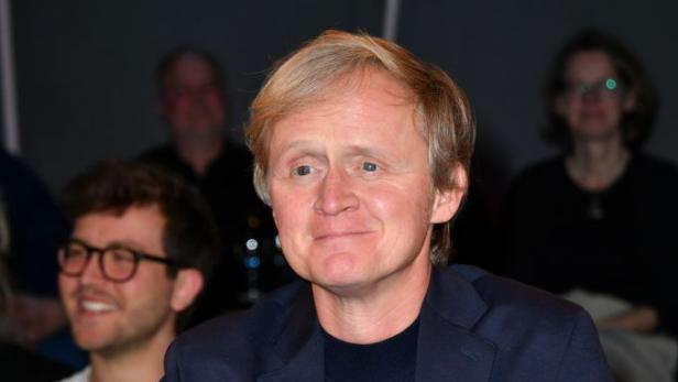 Pierre M. Krause sitzt in einer Gesprächsrunde, vor ihm stehen Blumen und ein Glas Wasser, im Hintergrund sind weitere Personen zu sehen.