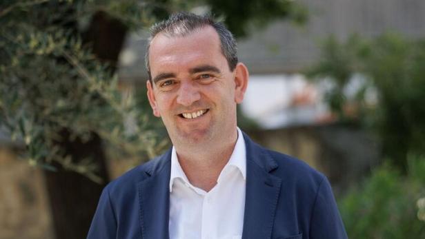 Ein Mann im blauen Anzug und weißem Hemd steht lächelnd im Freien vor grünen Pflanzen.