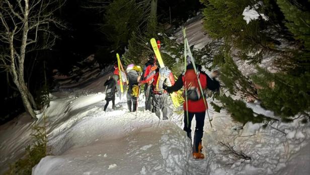 Mehrere Personen wandern nachts mit Skiern auf verschneitem Waldweg bergauf.