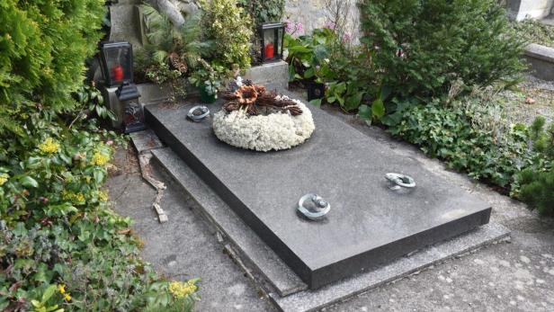 Grabstein mit Kranz aus weißen Blumen, drei kleinen Skulpturen und umgeben von grünen Pflanzen und Laternen mit roten Kerzen.