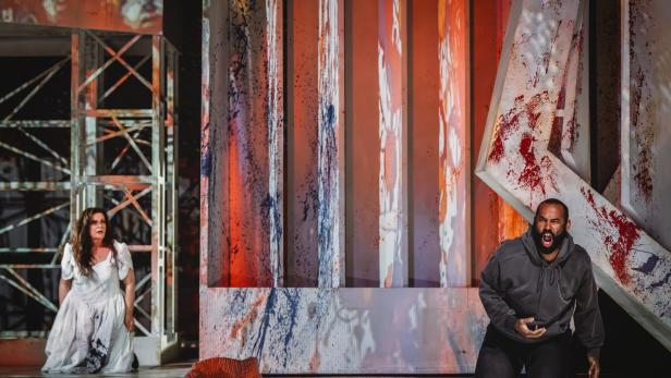 Eine Frau im weißen Kleid kniet links, ein Mann in dunkler Kleidung schreit kniend rechts vor blutverschmierten Kulissenteilen.