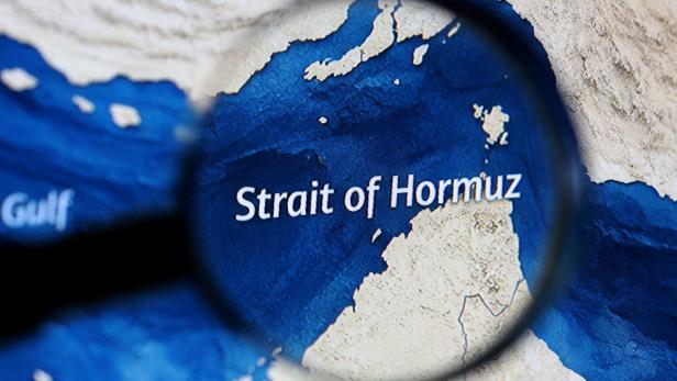 Eine Lupe vergrößert den Schriftzug „Strait of Hormuz“ auf einer Landkarte mit blauem Meer und beigen Küsten.