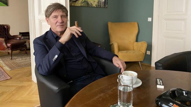 Ein Mann sitzt entspannt in einem Sessel, hält eine Zigarre, vor ihm stehen eine Tasse Kaffee und ein Glas Wasser auf dem Tisch.