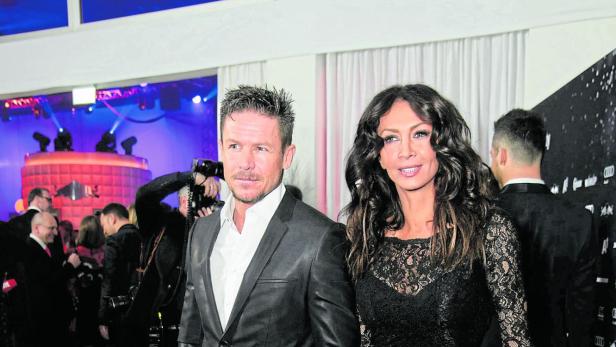 Ein elegant gekleidetes Paar hält Händchen auf einer festlichen Veranstaltung mit vielen Gästen im Hintergrund.